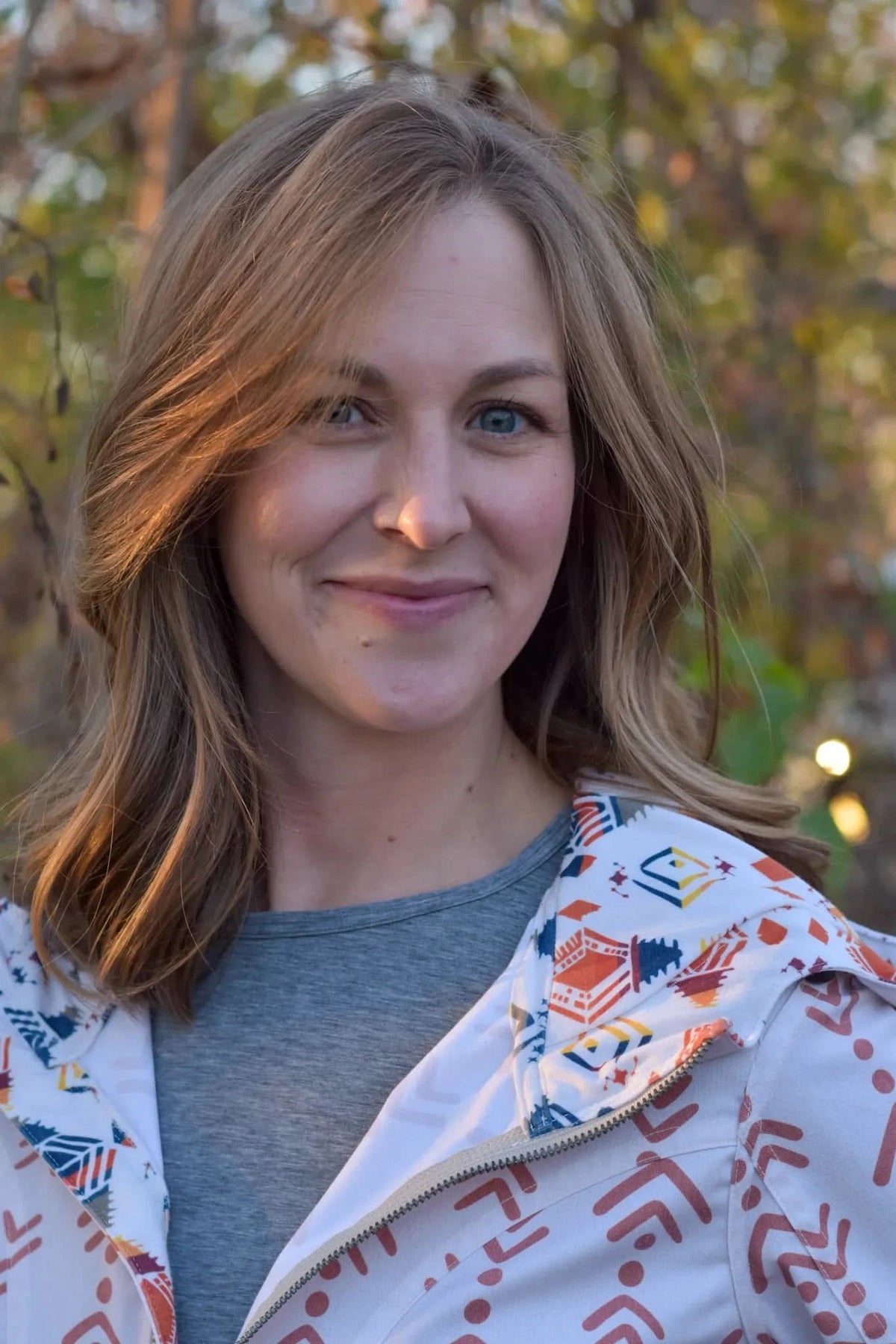 A woman smiles, wearing a white jacket with a colorful geometric pattern.