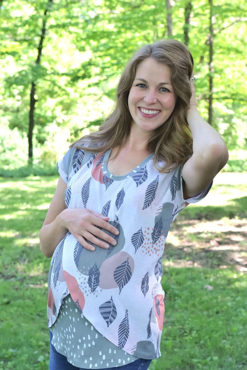A smiling pregnant woman wears a top with a mod leaf pattern in a park setting.