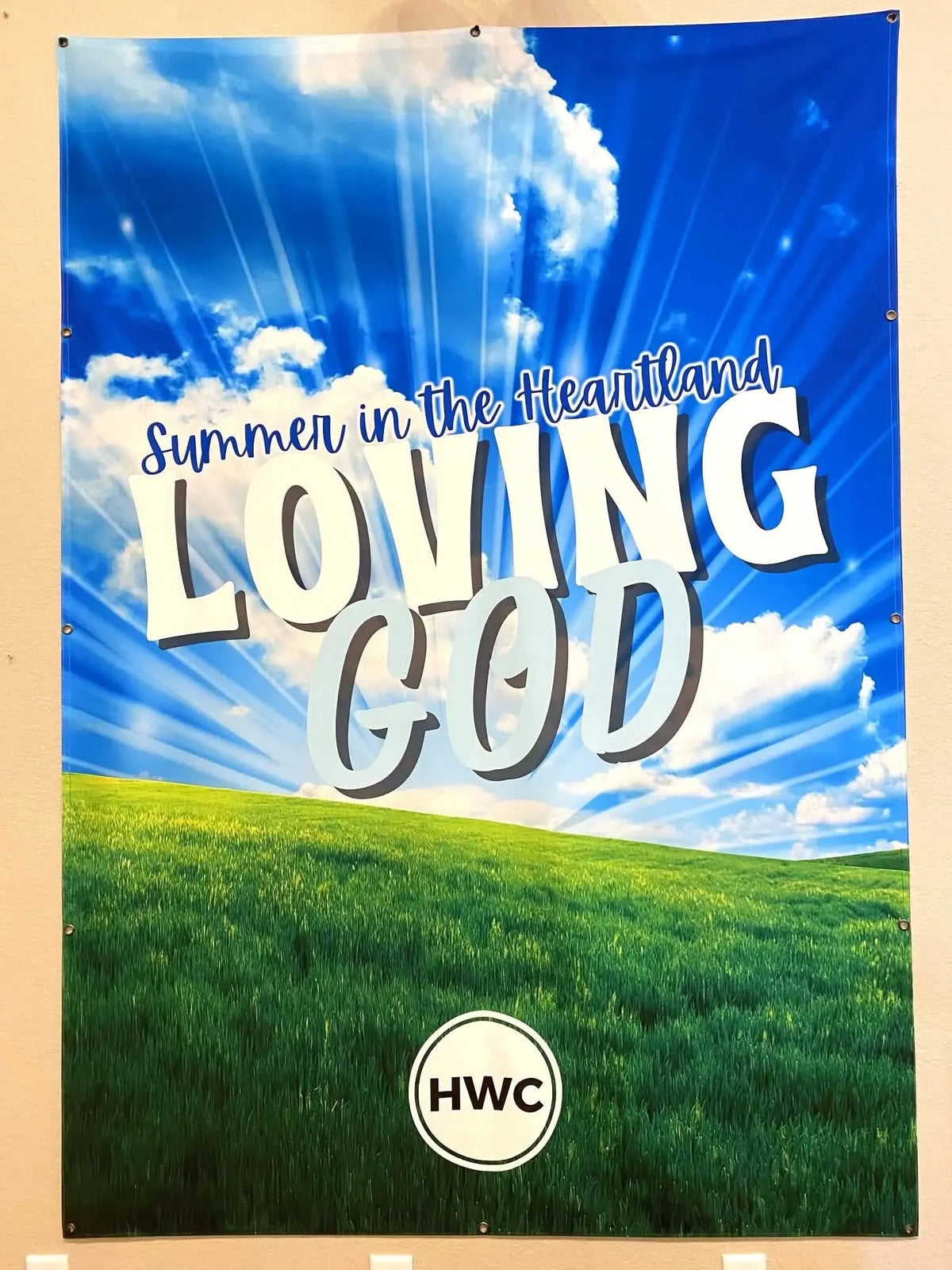 A banner with a blue sky, green field, and text "Summer in the Heartland Loving God HWC".