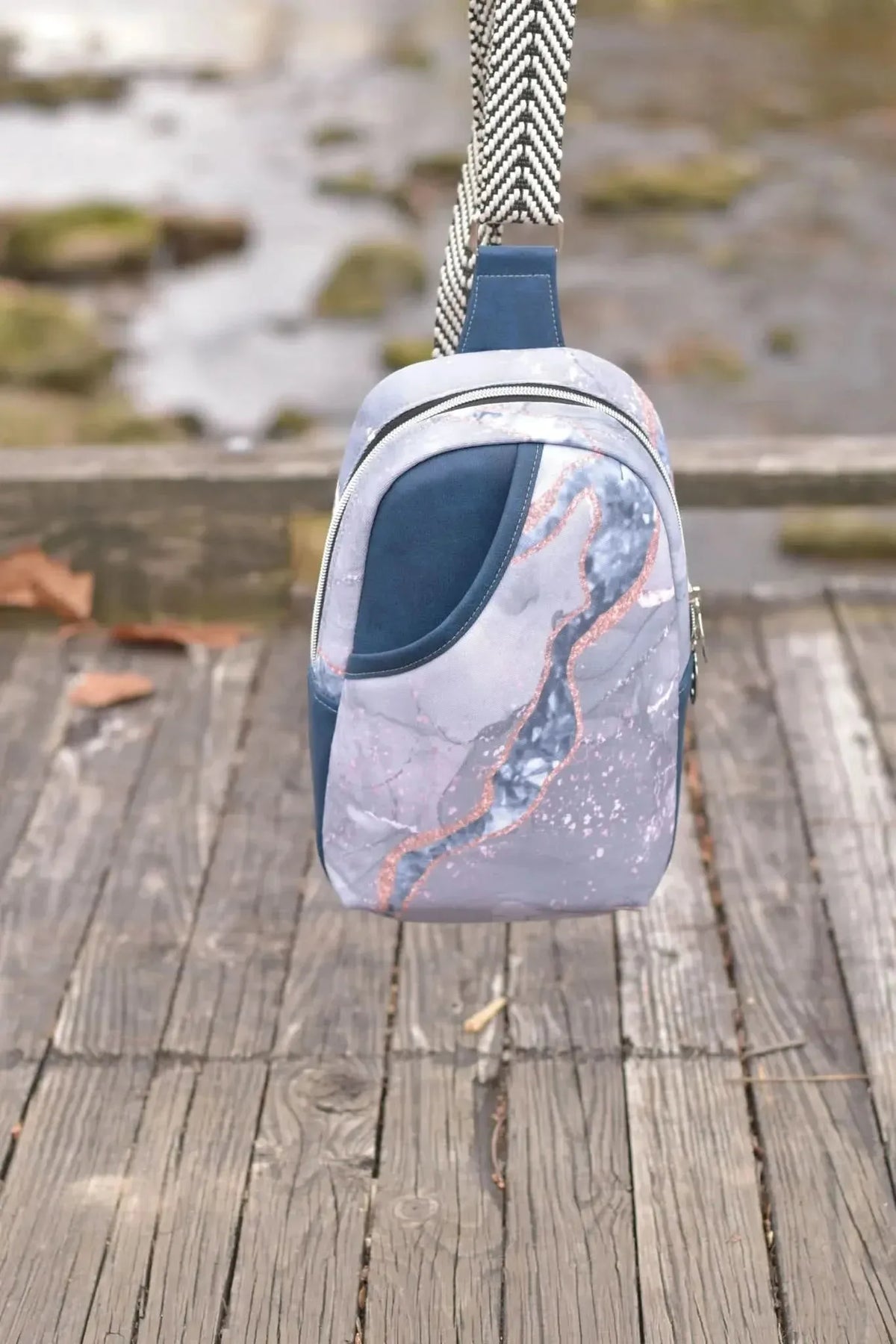 A blue and marble-patterned sling bag with a black and white strap hangs over a wooden railing.