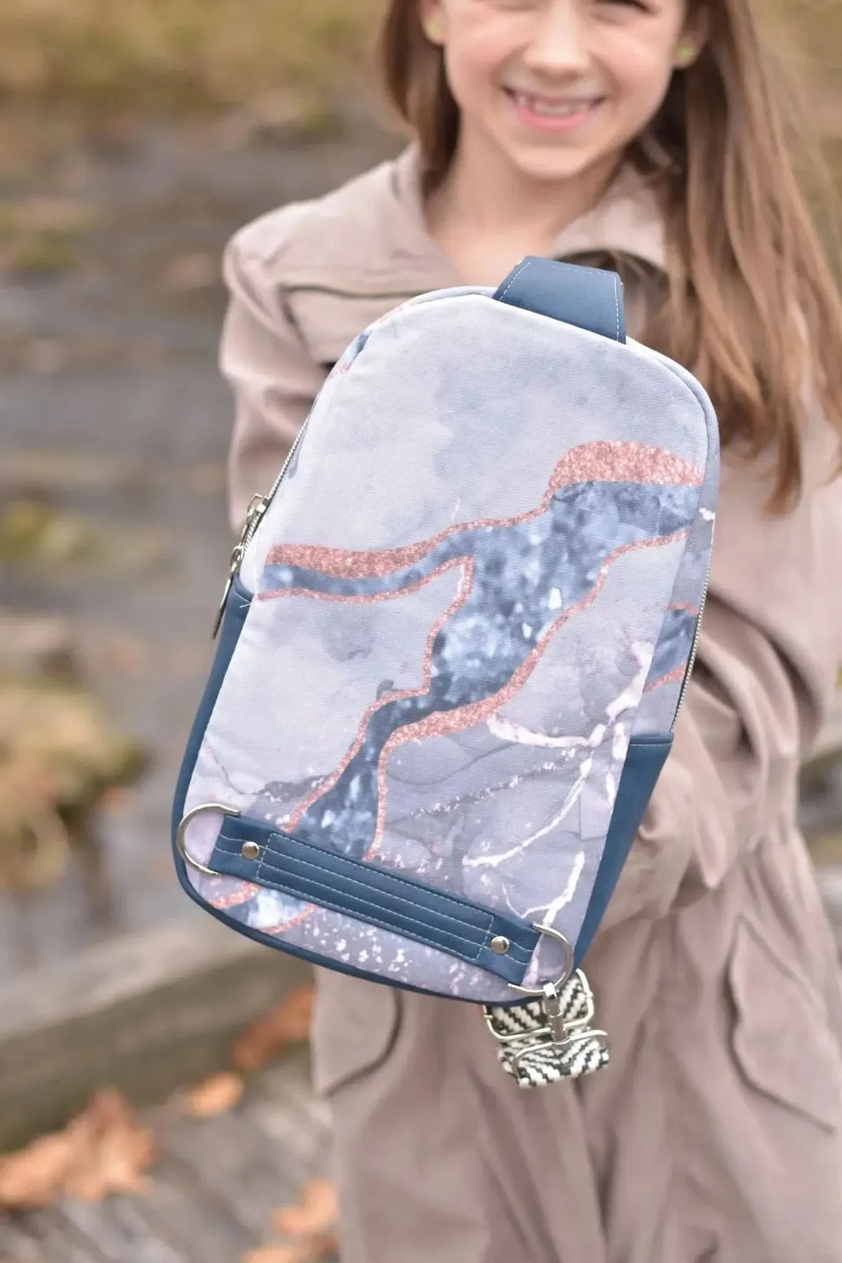 A young girl holds a blue marble-patterned sling bag with rose gold accents.