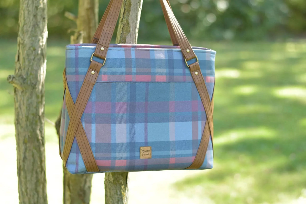 A plaid tote bag with brown leather straps hangs from a tree.