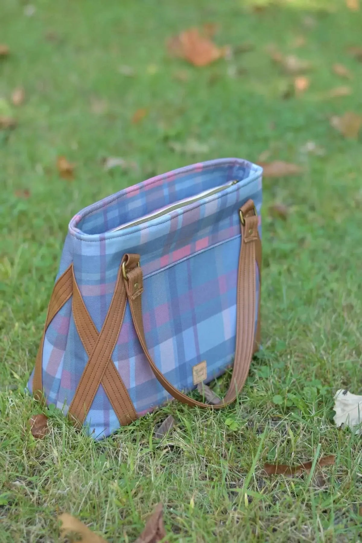 A blue and pink plaid tote bag with brown leather straps sits on green grass.