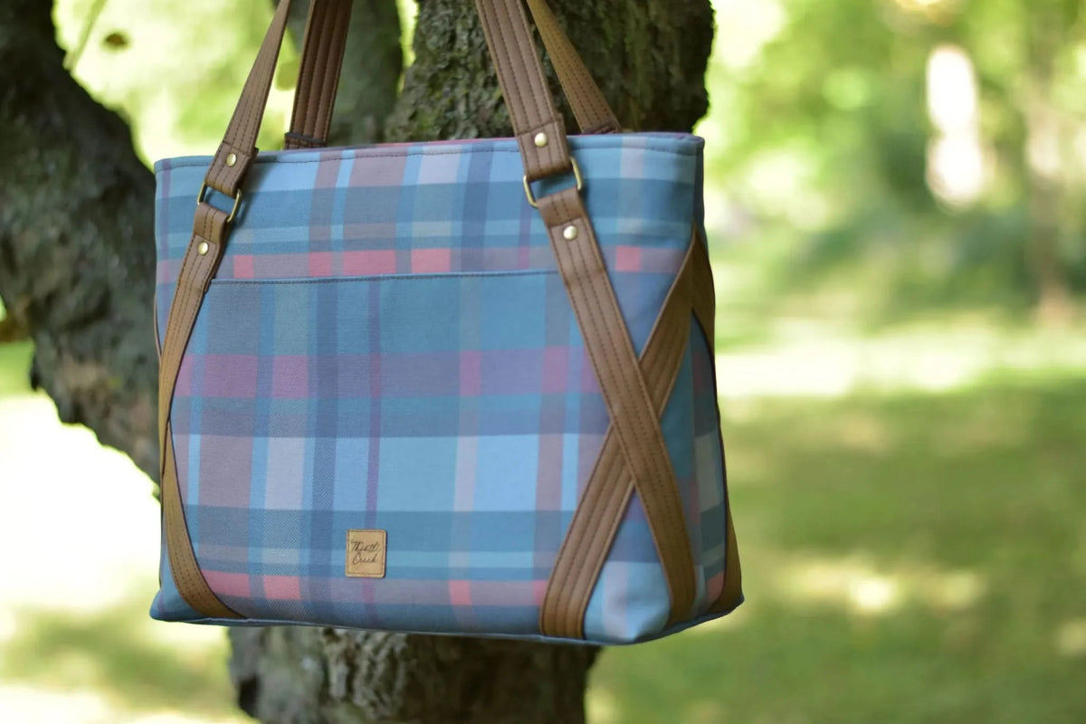 A plaid tote bag with brown leather straps hangs from a tree.
