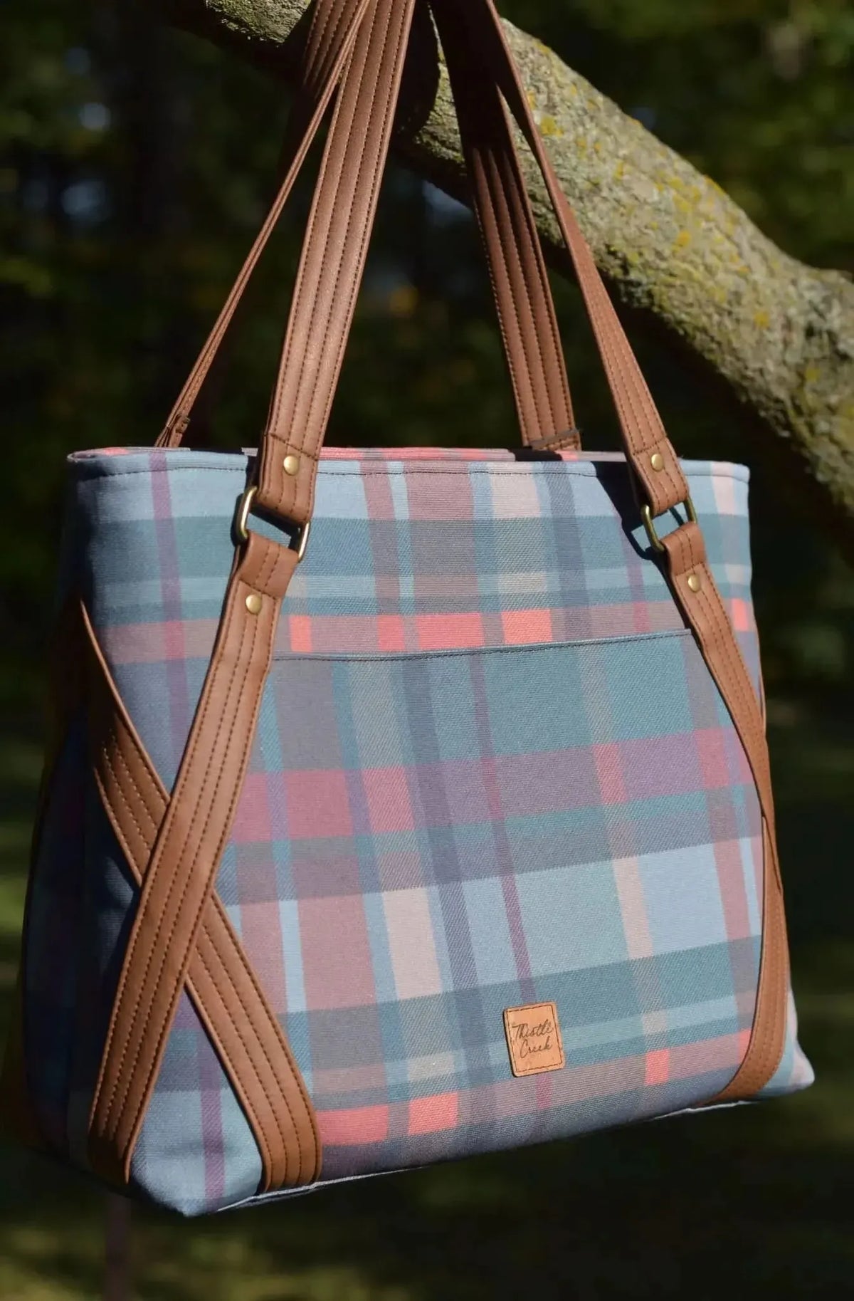A plaid tote bag with brown leather straps hangs from a tree branch.
