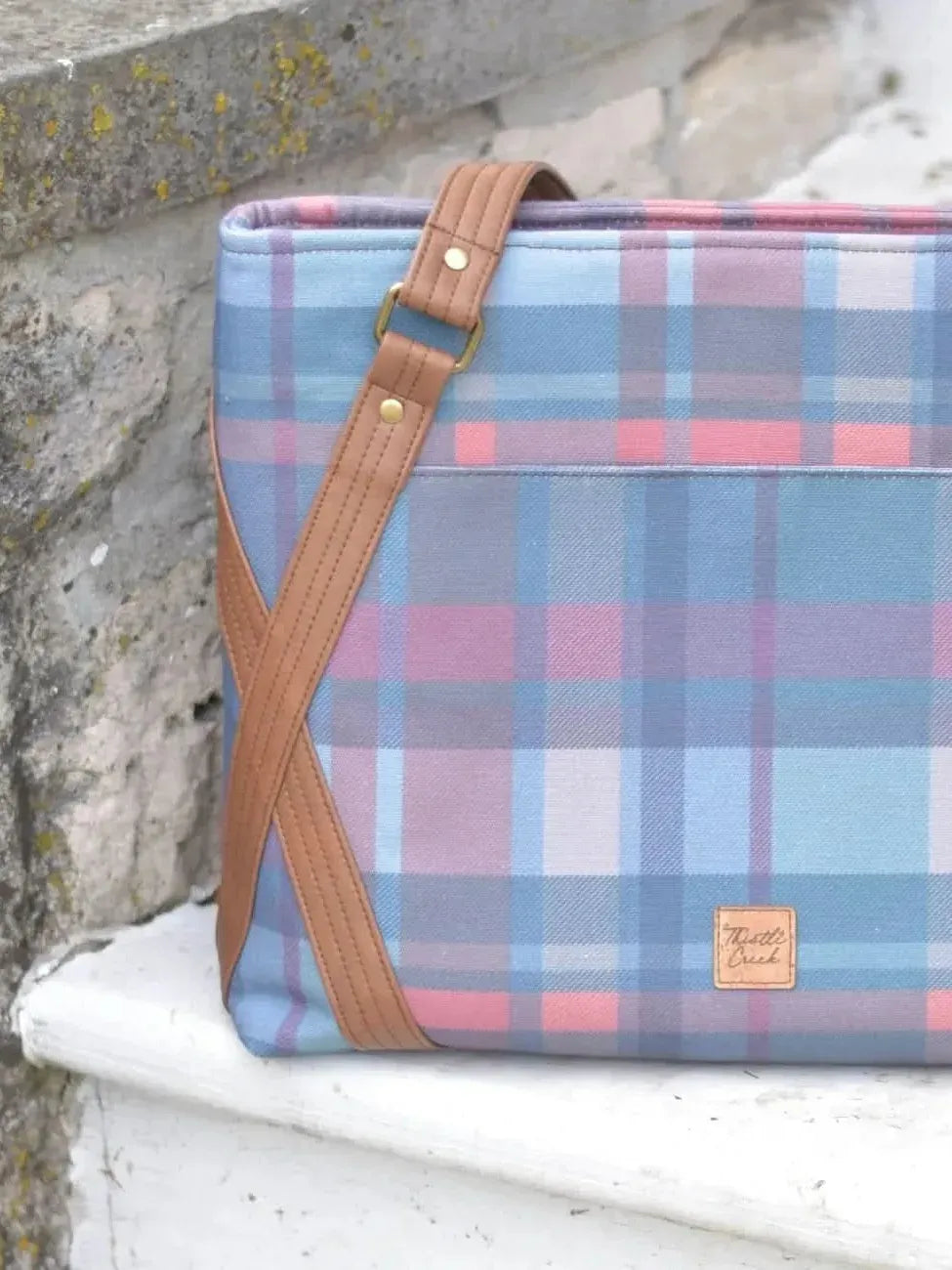 A plaid tote bag with brown leather straps sits on a stone ledge.