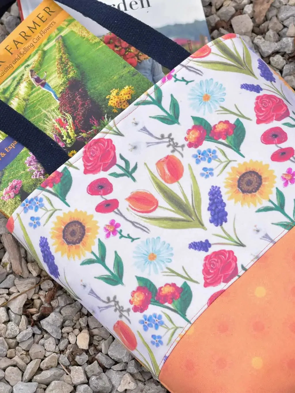 A floral tote bag with an orange bottom and two books on a bed of gravel.
