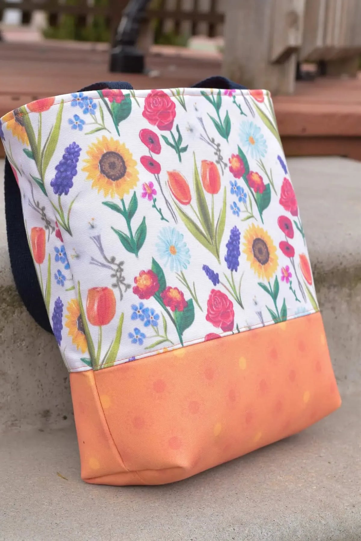A tote bag with a colorful garden flower pattern and a solid orange bottom.