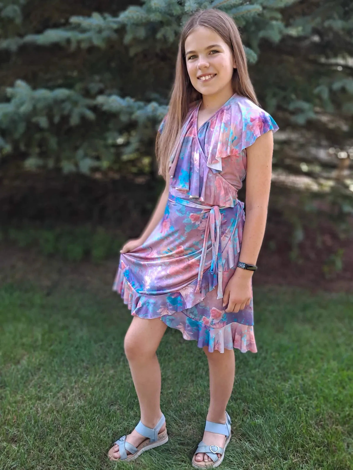 A young girl wears a purple floral wrap dress and light blue sandals.