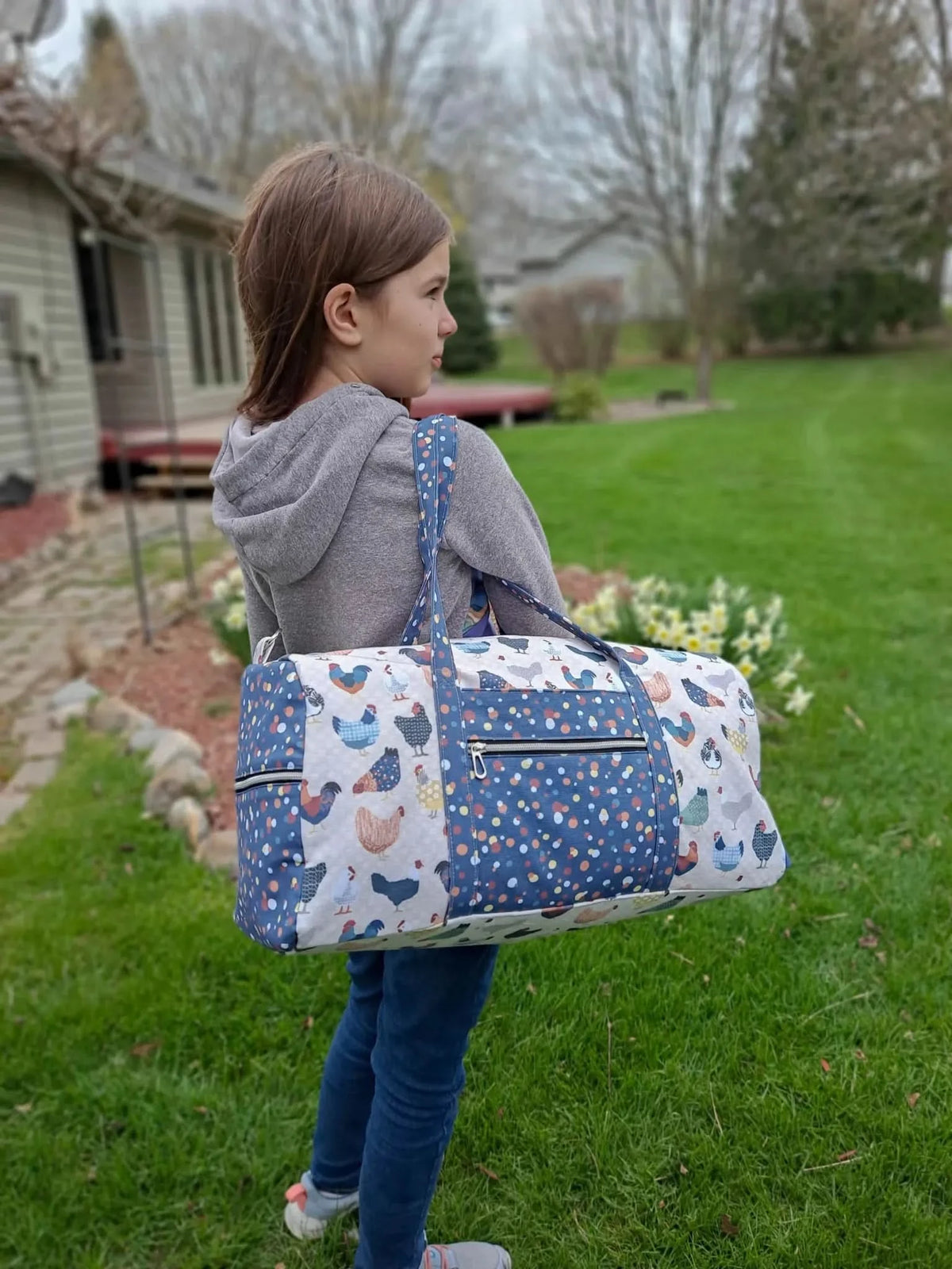 A young girl carries a duffel bag with a patchwork chicken and polka dot pattern.