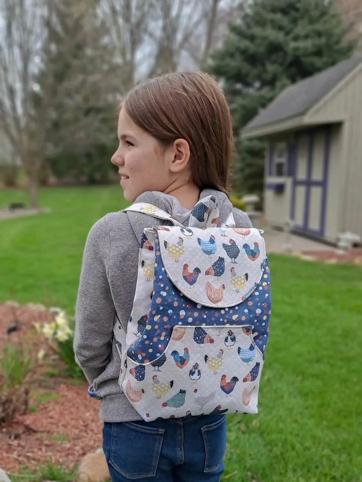 A young girl wears a backpack with a patchwork chicken pattern.