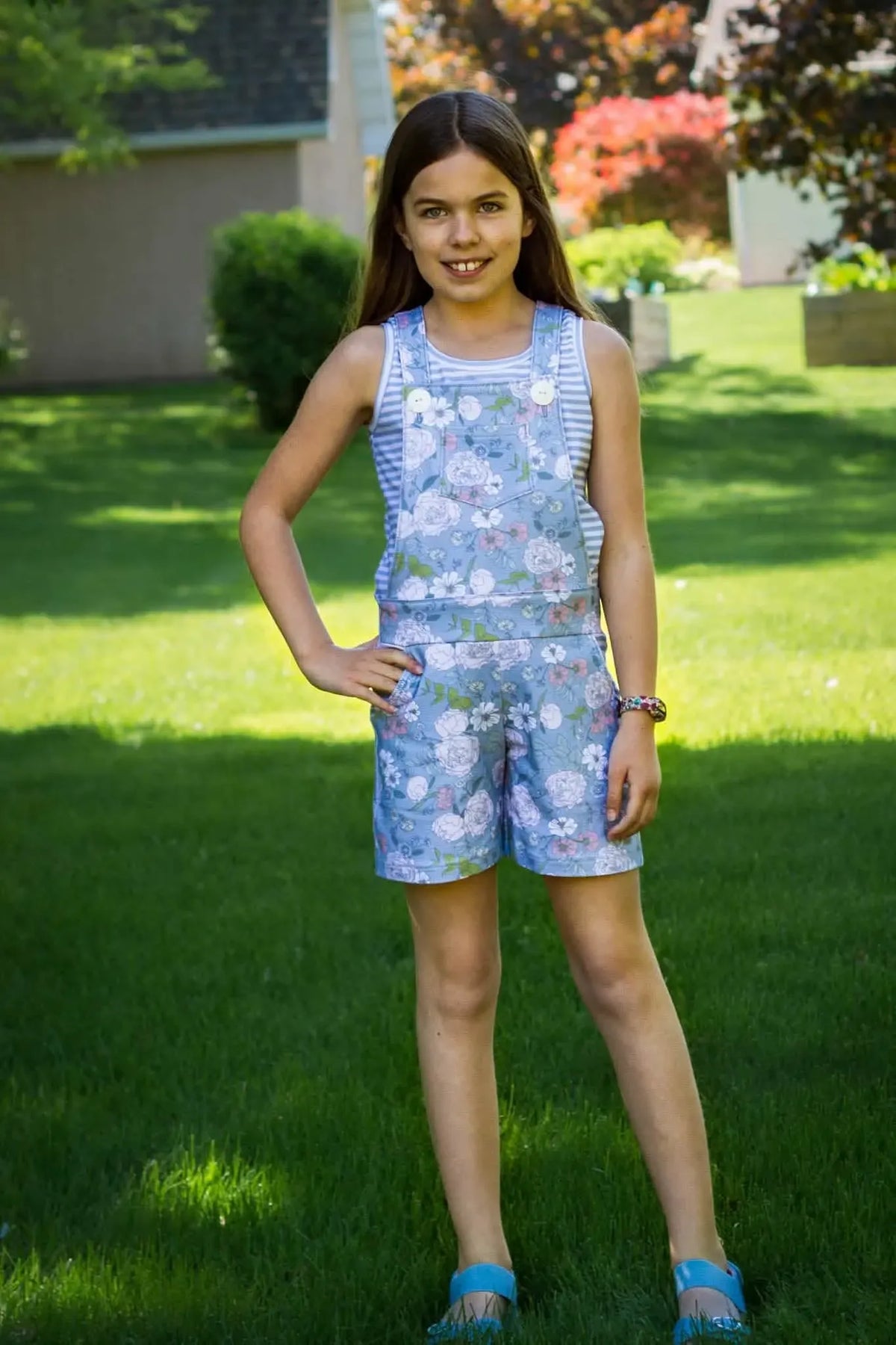 A young girl wears a dusty blue floral romper with a striped shirt underneath.