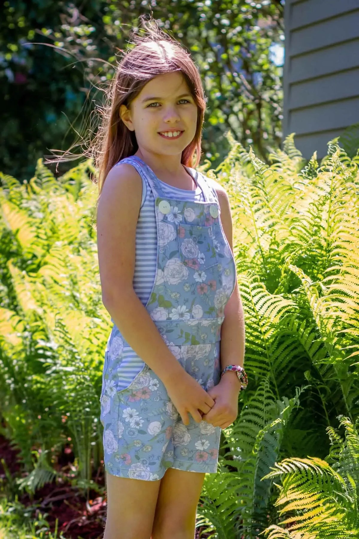 A young girl wears a dusty blue floral overall dress over a striped shirt.