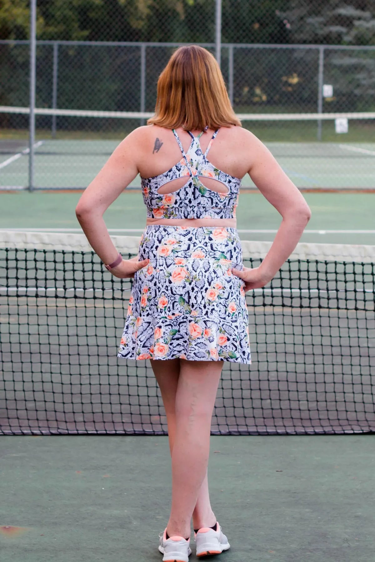 A woman from behind wears a floral snake print athletic top and skirt on a tennis court.