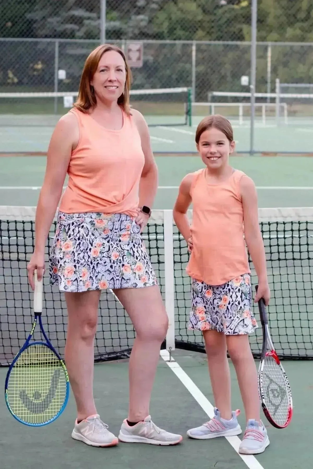 Mother and daughter in matching orange floral snake print skirts and tanks on tennis court.