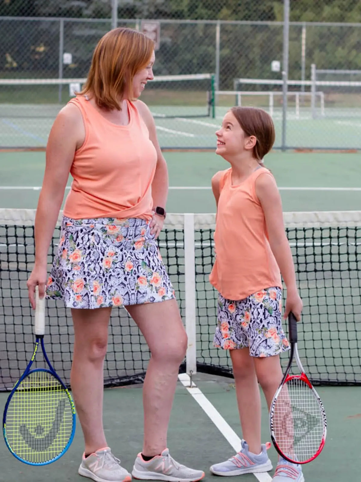 A woman and girl wear matching floral snake print athletic skirts on a tennis court.