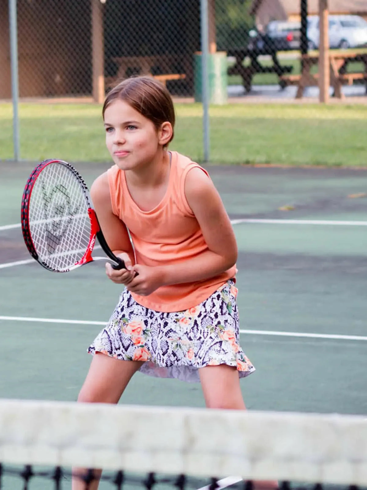 A young girl in a floral snake print skirt plays tennis on a green court.