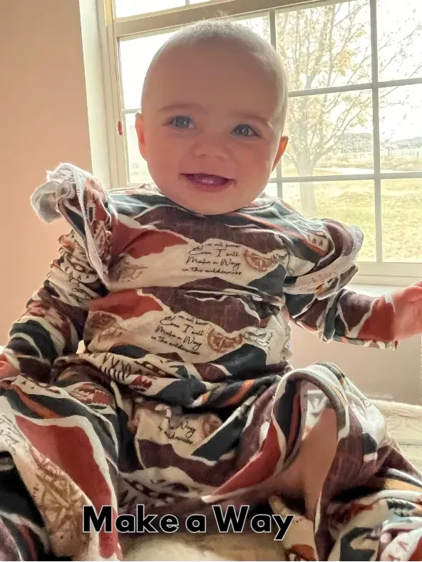 A baby in a brown, white, and black patterned French terry outfit smiles at the camera.