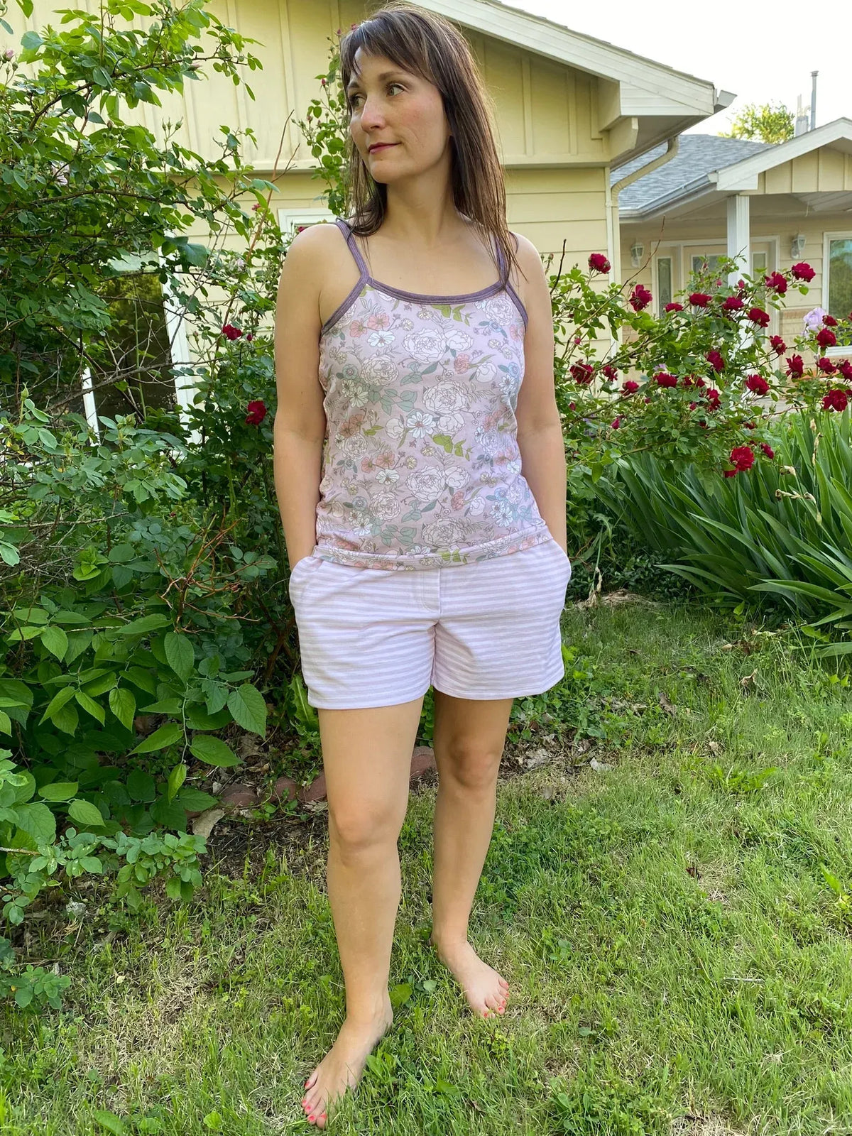 Woman modeling a mauve striped tank top and shorts made from stretchy fabric, standing barefoot in a garden.