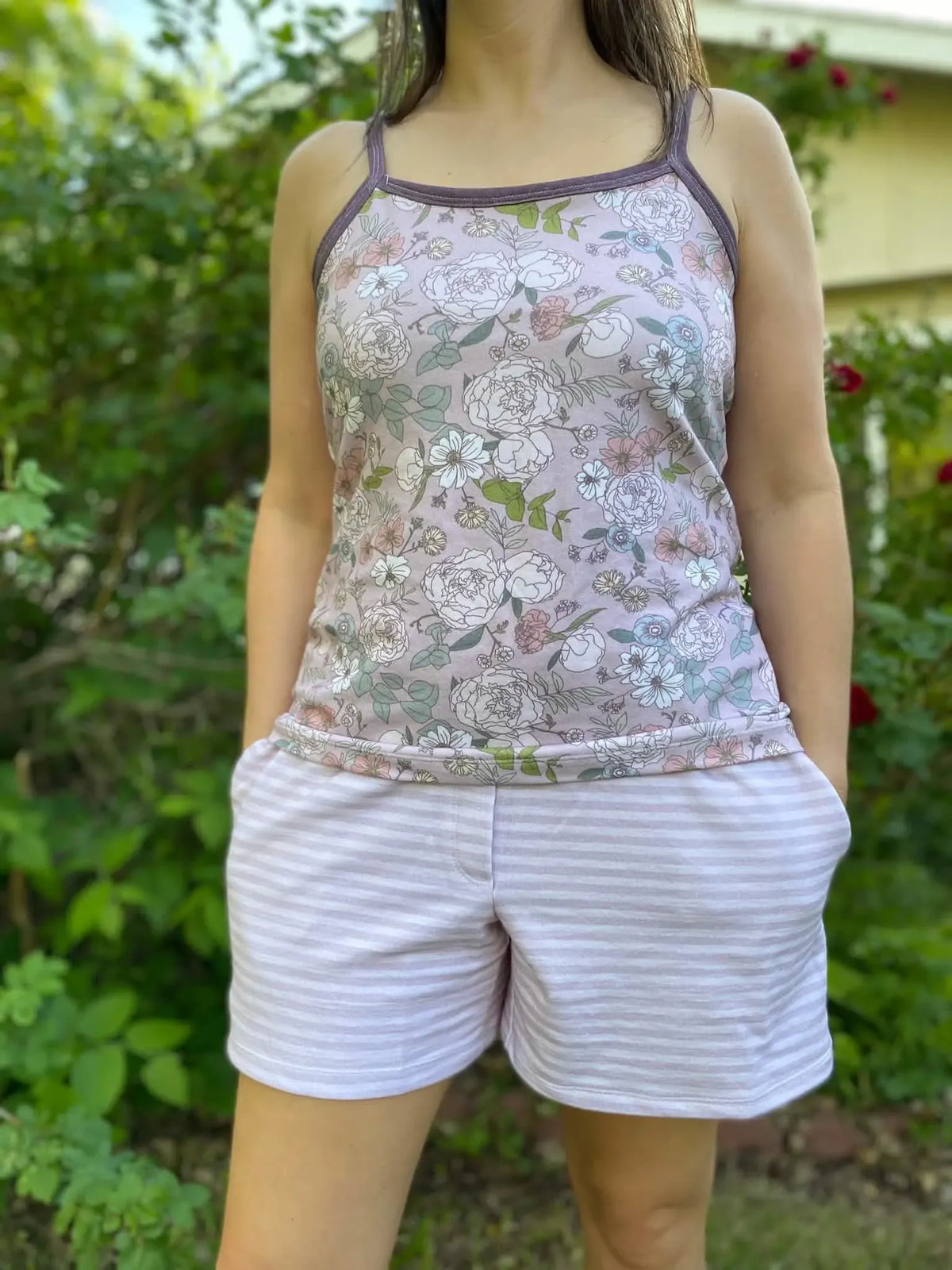 A person wearing a floral tank top and striped shorts stands in a garden.