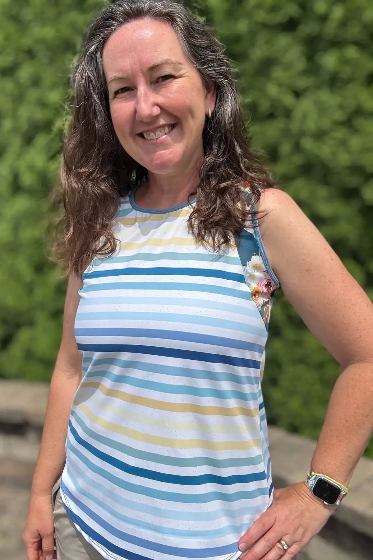 A woman smiles, wearing a striped tank top with blue, yellow, and white colors.