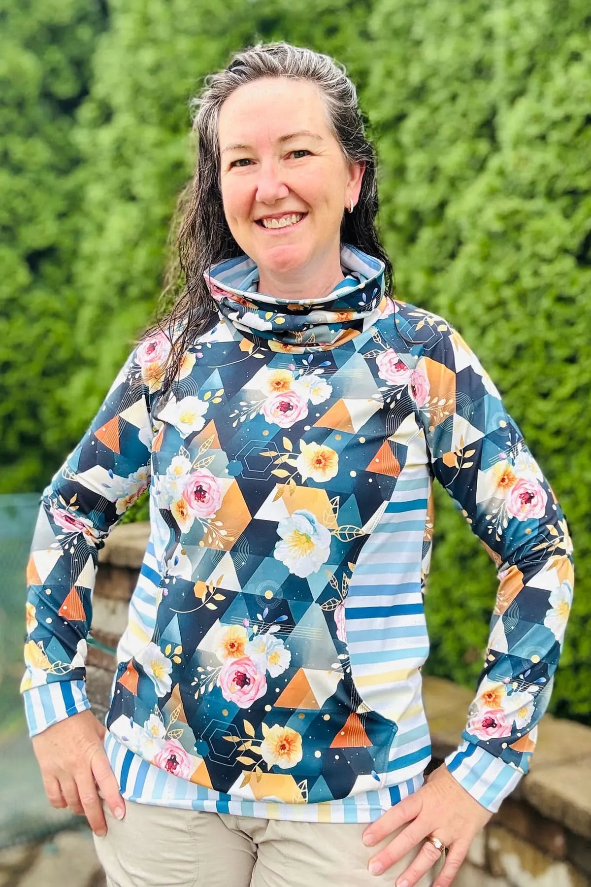 A woman smiles wearing a long-sleeved top with a floral and geometric pattern.