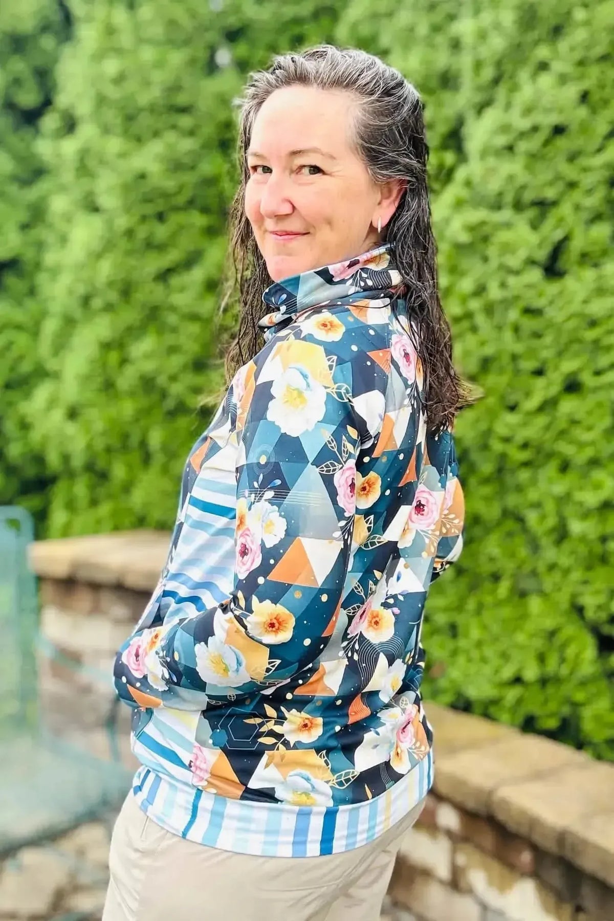 Woman wearing a dark teal geometric floral print hoodie with side pockets, viewed from the back against a green hedge.