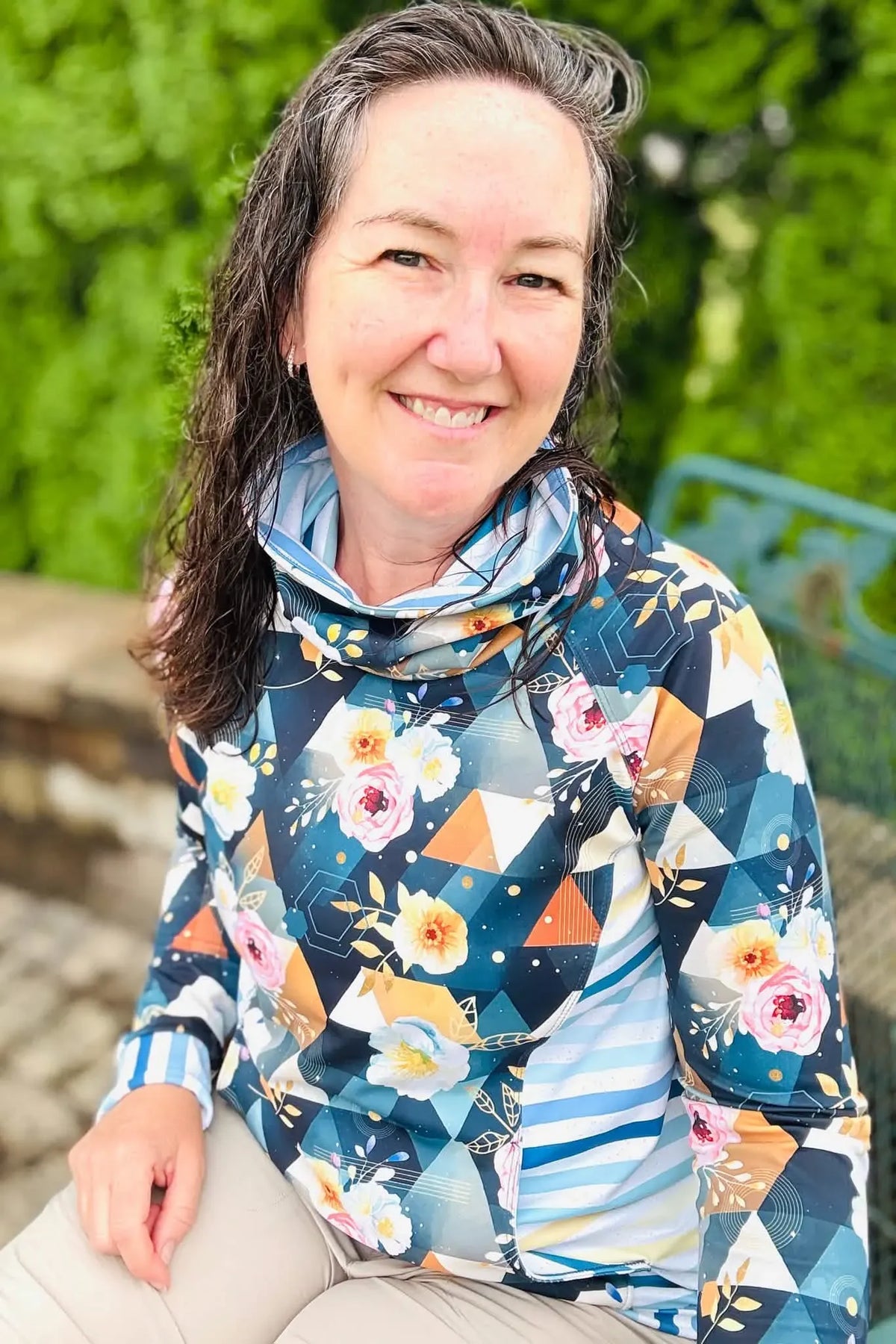 A woman smiles, wearing a long-sleeved top with a floral and geometric pattern.