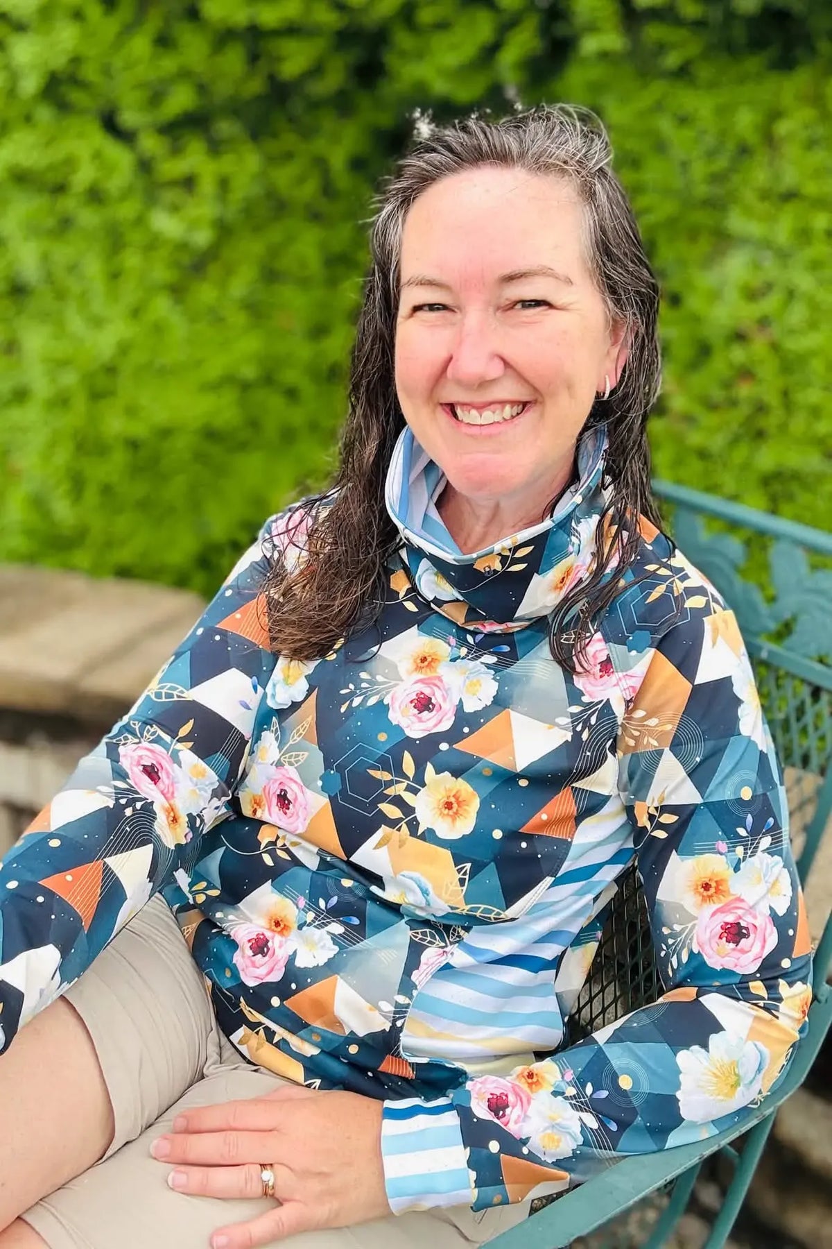 A woman smiles, wearing a long-sleeved top with a floral and geometric pattern.