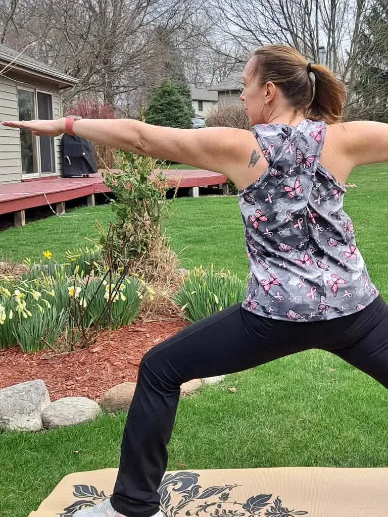 A woman in a gray tank top with pink butterflies and black pants practices yoga outdoors.