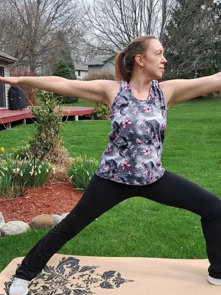 Woman wearing tank top with pink ribbons and butterfly print fabric in warrior yoga pose on mat.