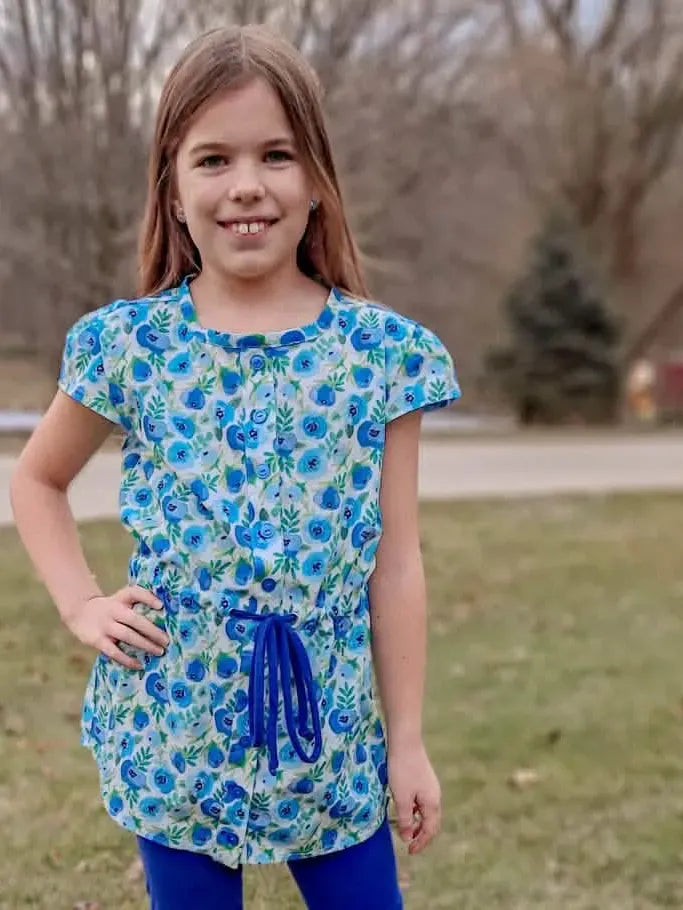 A young girl smiles, wearing a blue floral top with a drawstring waist and blue leggings.