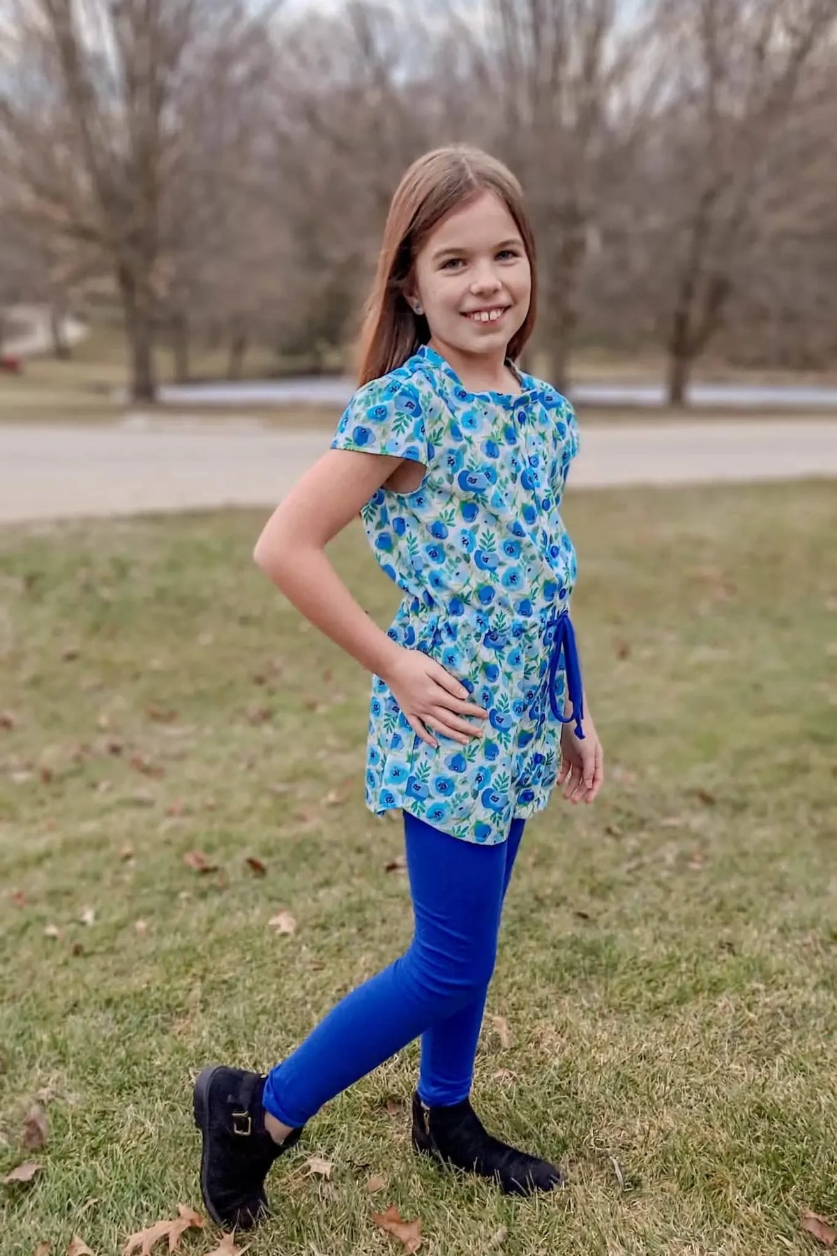 A young girl smiles, wearing a blue buttercup patterned top and blue leggings.