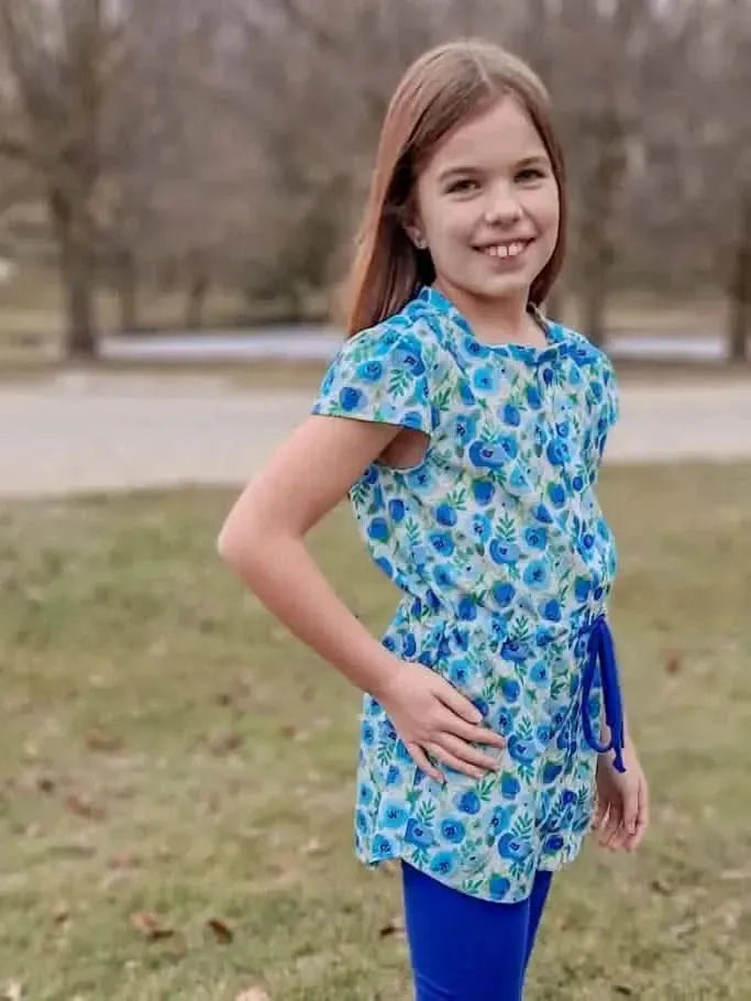 A young girl wears a blue buttercups floral print top and blue leggings.