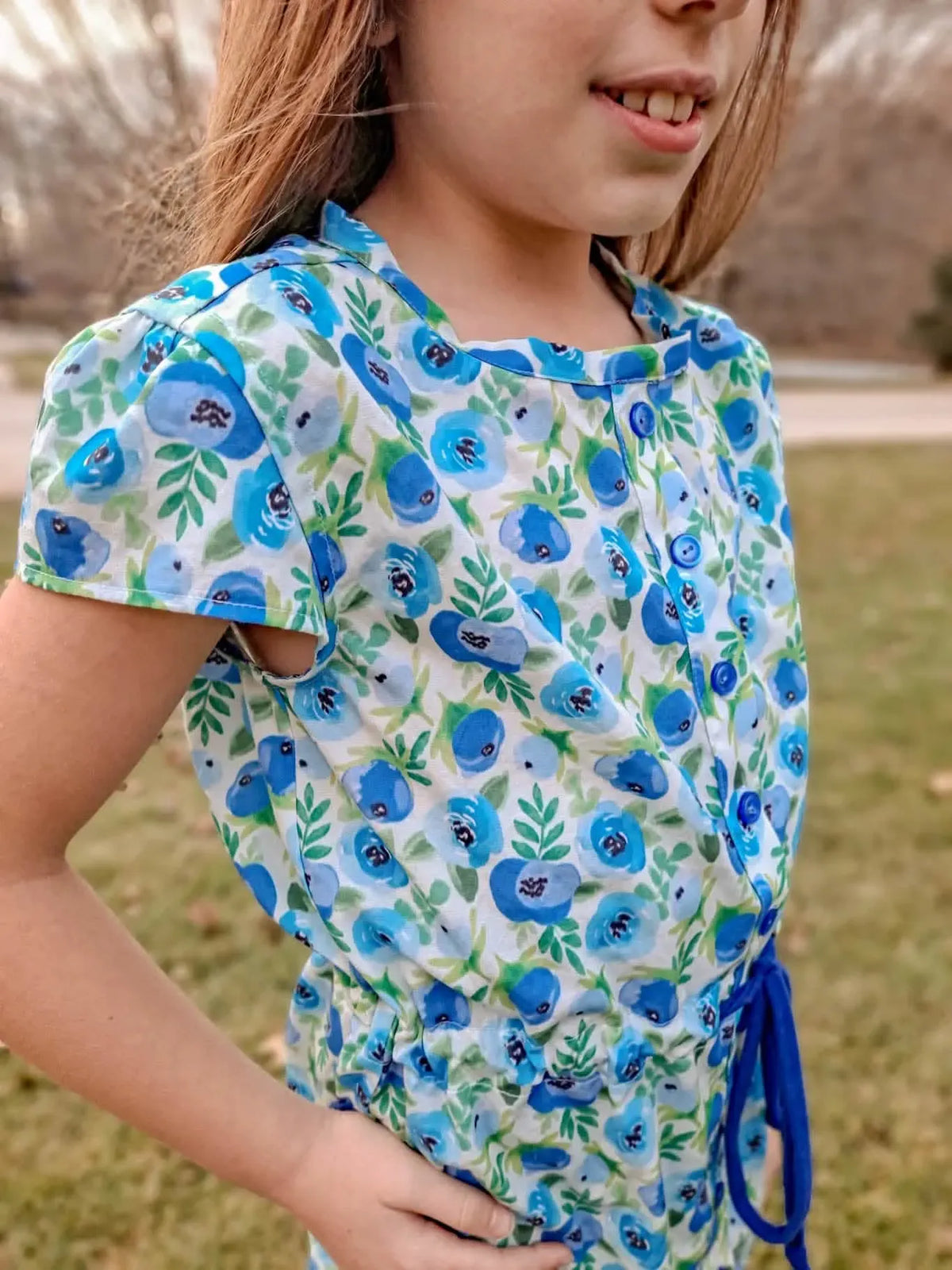 A child wears a blue buttercup floral print dress with blue buttons and a drawstring.