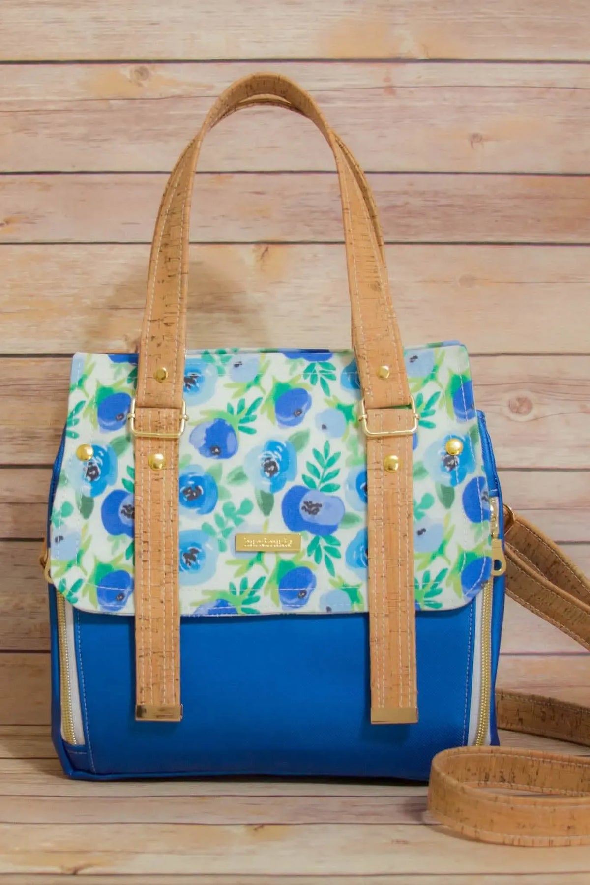 A blue and white floral handbag with cork straps sits on a wooden surface.
