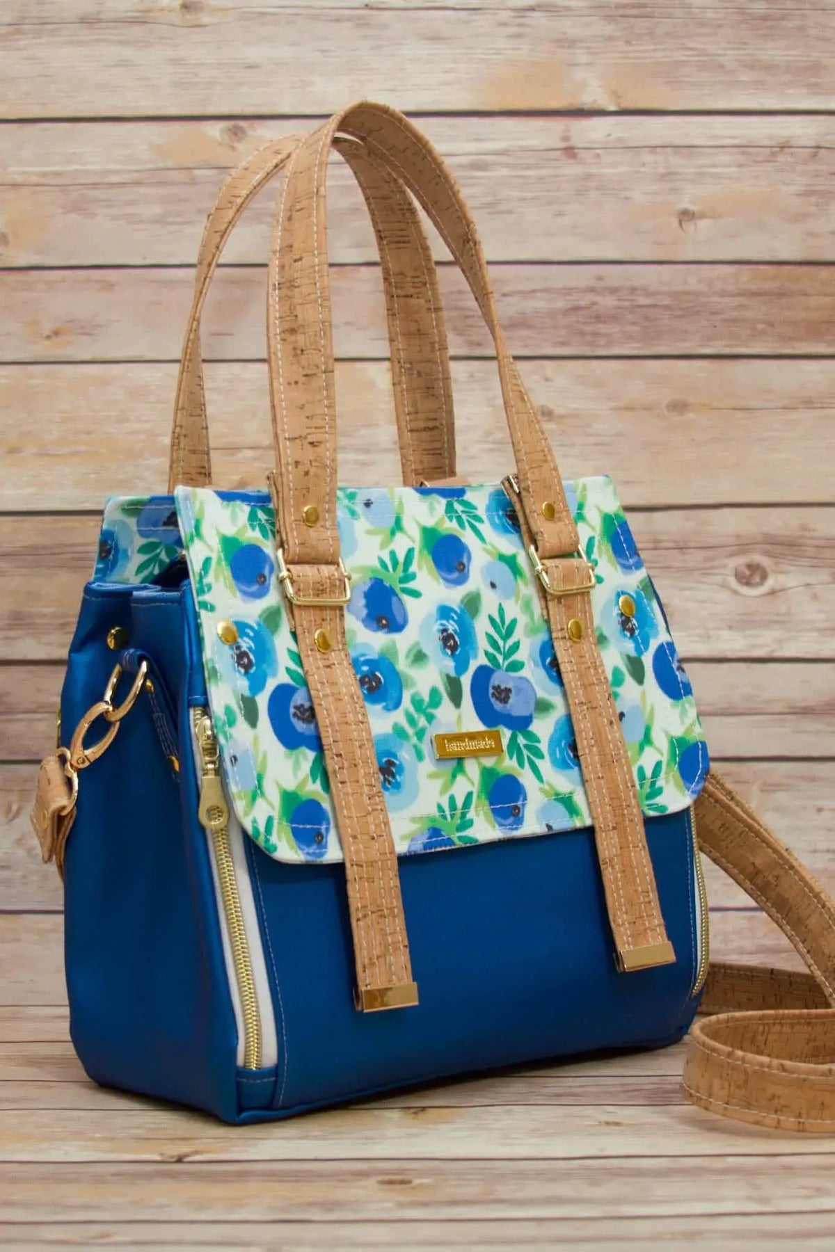A blue handbag with a floral flap and cork-like straps sits on a wooden surface.