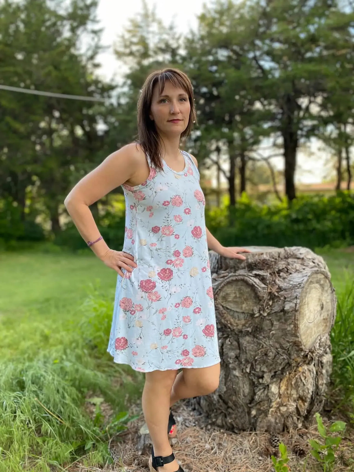 A woman in a light blue floral dress stands next to a tree stump outdoors.