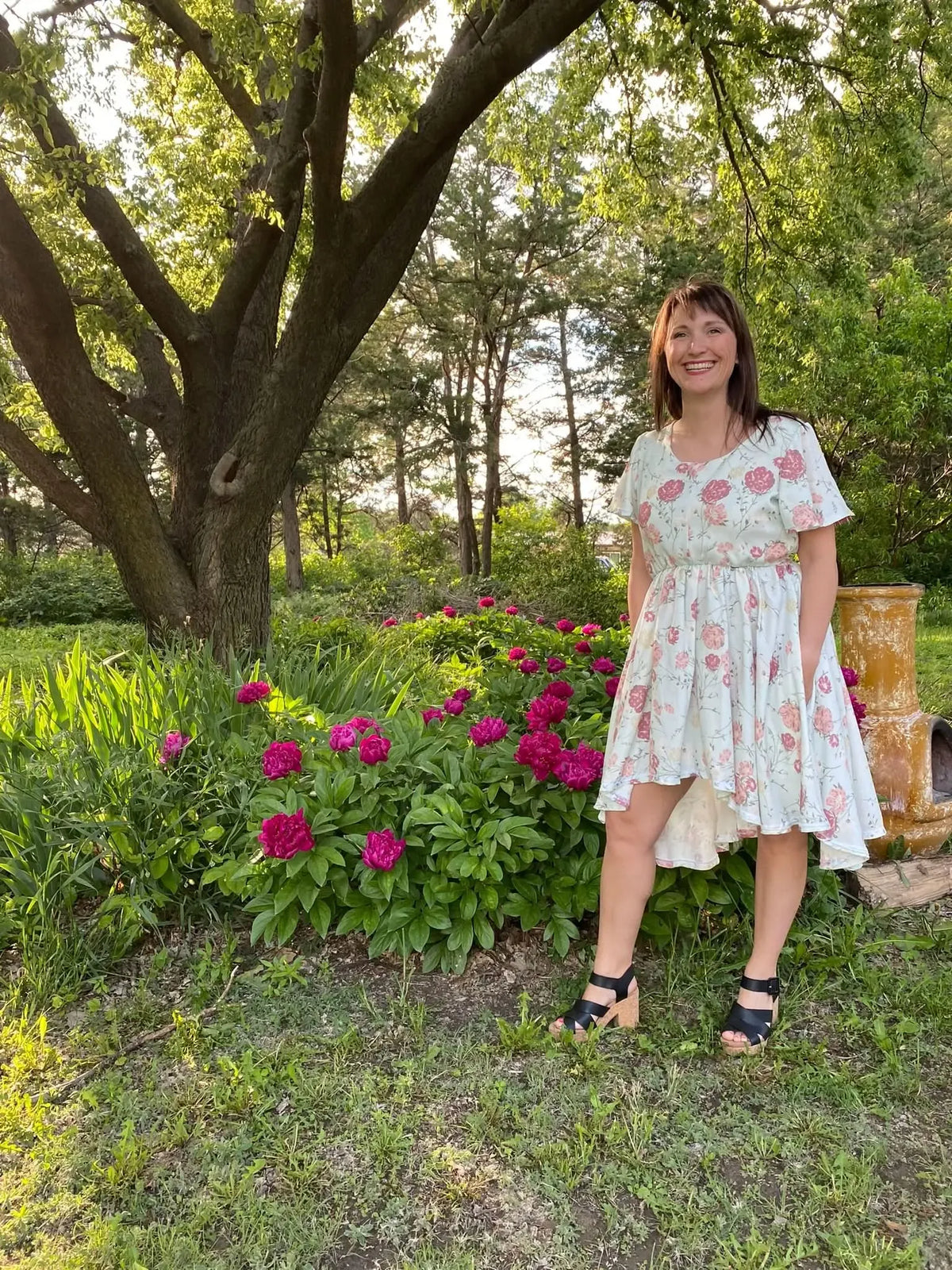 A woman in a floral dress stands in a garden with pink peonies and a large tree.