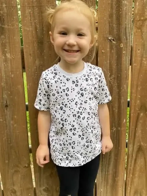 A smiling child wears a white shirt with a black leopard print.