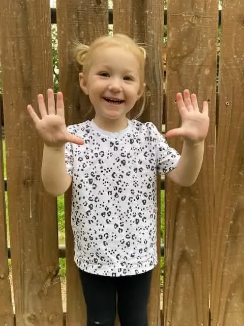 A smiling child in a white shirt with black leopard print stands in front of a fence.