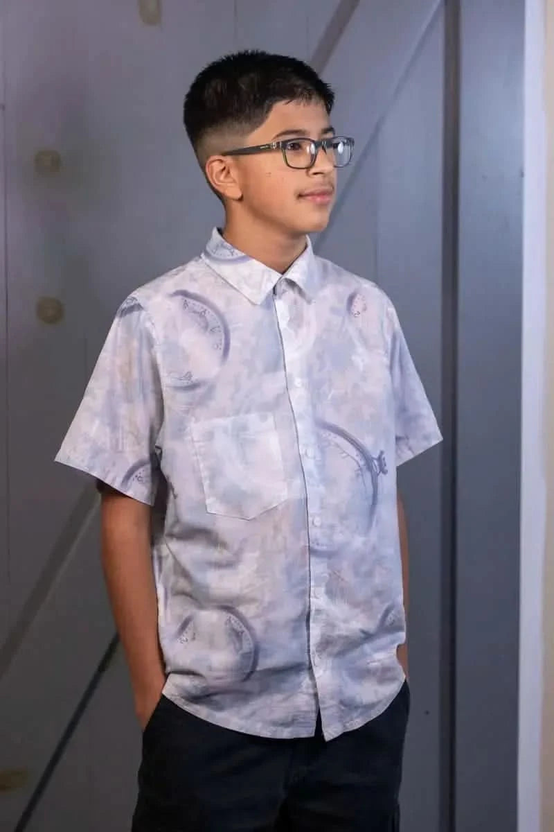 A boy in a blue patterned short-sleeved button-up shirt and dark pants.