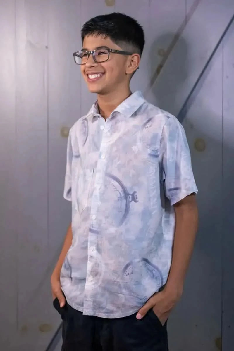 A boy in a blue short-sleeved button-up shirt with a clock pattern and dark shorts.
