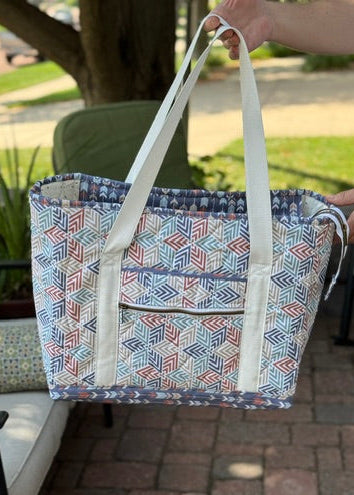 A person holds up a large tote bag with a blue, red, and white geometric pattern, set against an outdoor patio background with a brick floor and patio furniture.