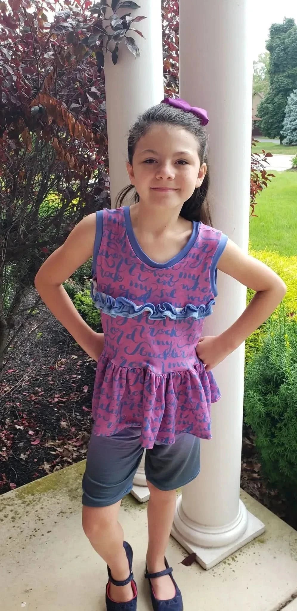 A young girl wears a pink and blue ruffled top with dark shorts and blue shoes.