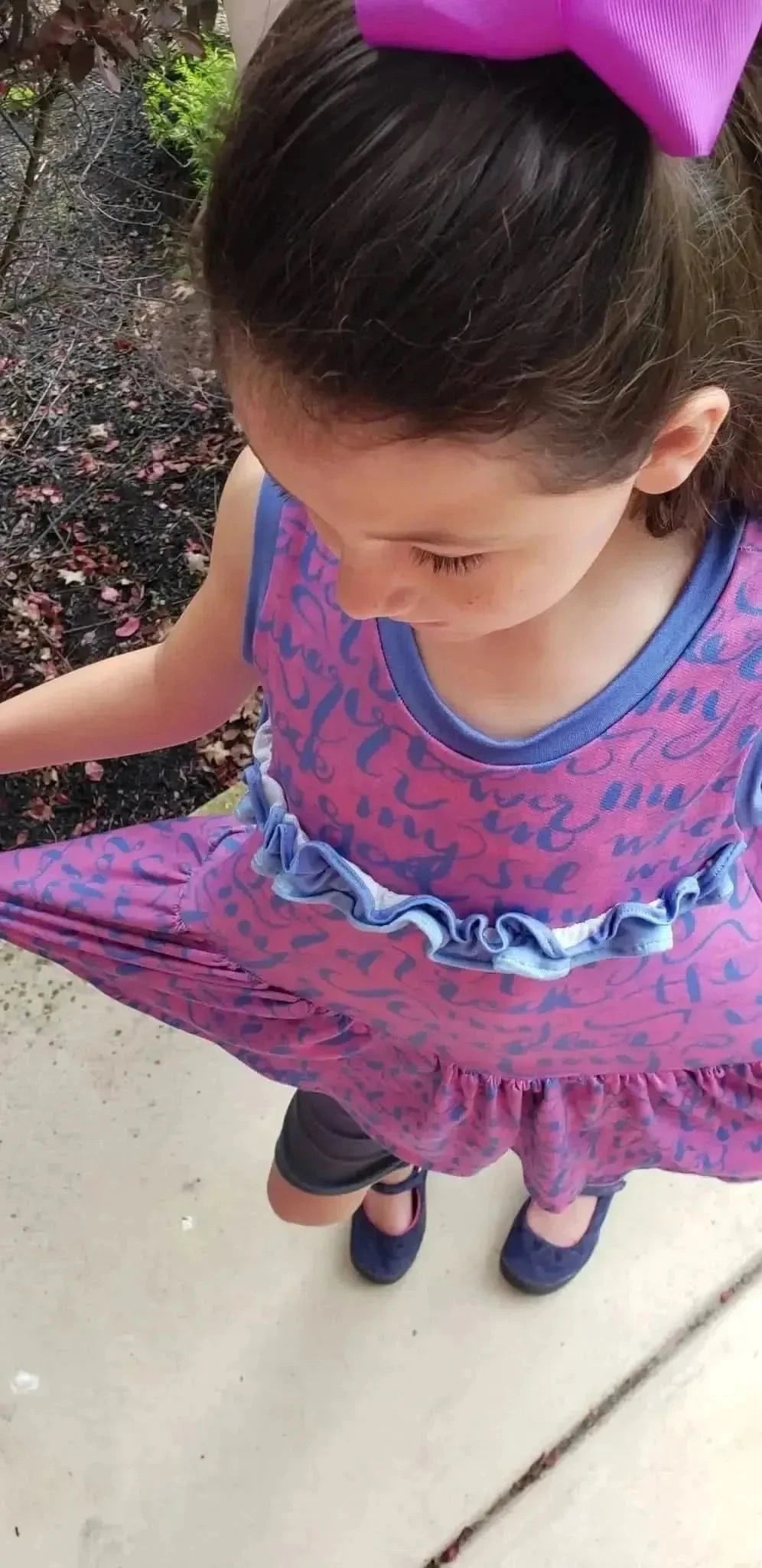 A young girl wears a pink dress with blue script and a purple bow in her hair.
