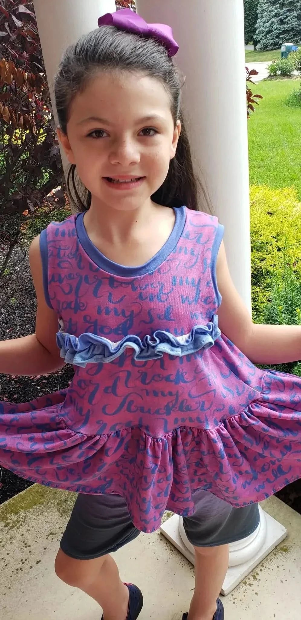 A young girl smiles, wearing a pink and blue patterned dress with ruffles.