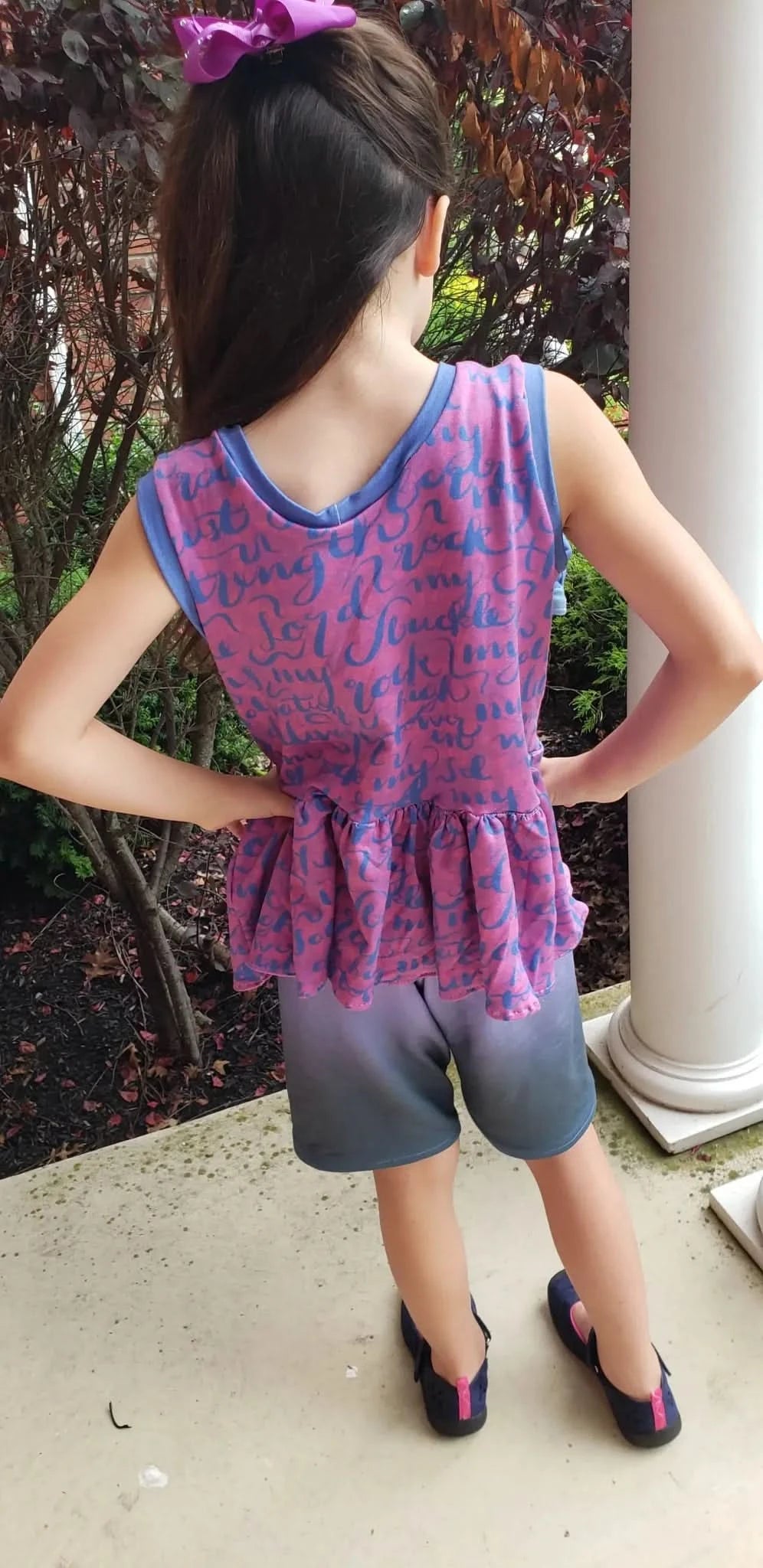 A child from behind wears a pink and blue top with gray shorts and blue shoes.