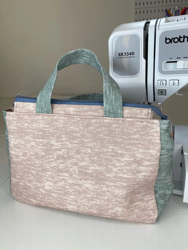 A light pink and green fabric bag with a zipper and handles sits in front of a sewing machine.
