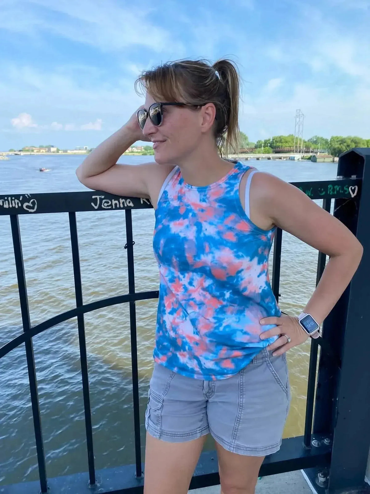 A woman wears a blue and pink tie-dye tank top and gray shorts by a river.